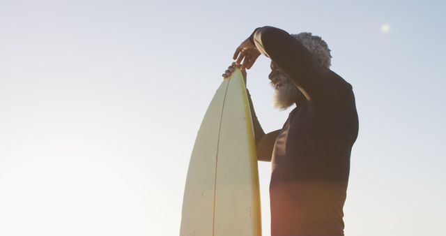 Senior man waxing a yellow surfboard in bright sunshine at the beach. Great for illustrating active lifestyles, mature athletes, fitness, peaceful beach moments, and outdoor hobbies. Can be used in advertisements for surf gear, retirement activities, beach resorts, fitness campaigns, and wellness promotions.