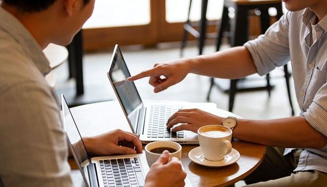 Colleagues collaborating over laptops and coffee in sunlit cafe, pointing at screen, teamwork