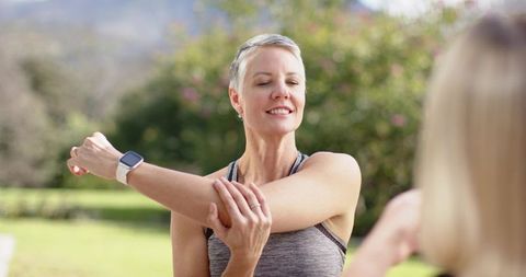 Mature woman stretching with workout partner in park wearing smartwatch and activewear