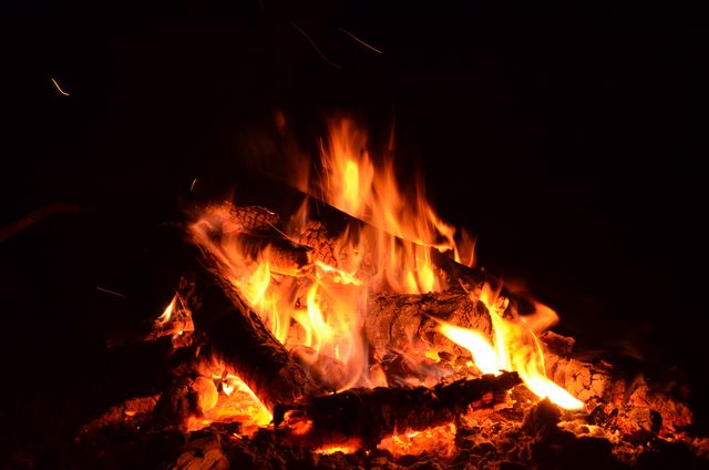 Burning campfire emits glowing flames and sparks in dark night. Ideal for projects on outdoor adventures, camping trips, summer nights, and wilderness survival. Shows warmth, gathering around fire, rustic experience, and connection with nature.