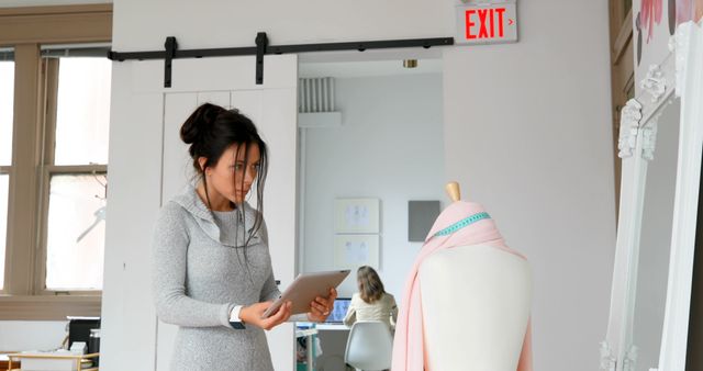 The image shows a female fashion designer using a tablet while examining a mannequin in a modern studio. She seems to be assessing or choosing fabric draped over the mannequin. In the background, another person sits and works at a desk. This image is suitable for content related to fashion design, creative professionals, modern workspaces, and the integration of technology in creative industries.