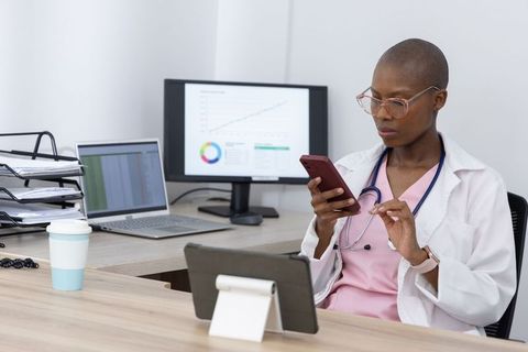 African american female doctor reviewing smartphone at desk with laptop and charts