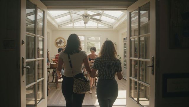 Two women walking through french doors into sunlit conservatory gathering at dining table