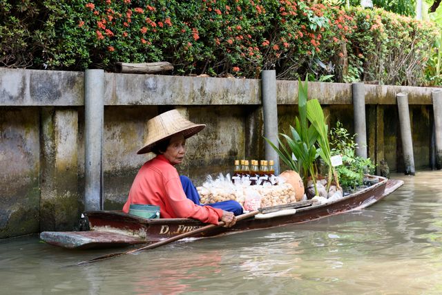 This depicts a vendor in a traditional wooden boat selling local goods on a river, representing the unique marketplace culture. Ideal for use in cultural articles, travel blogs, or promotional material for tourism in Thailand. It highlights traditional market practices in a natural river setting.