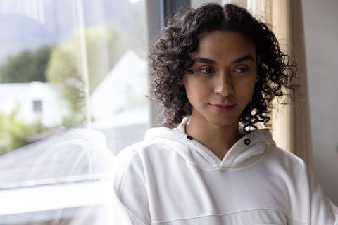 Androgynous non-binary adult standing by window wearing off-white hoodie with reflection