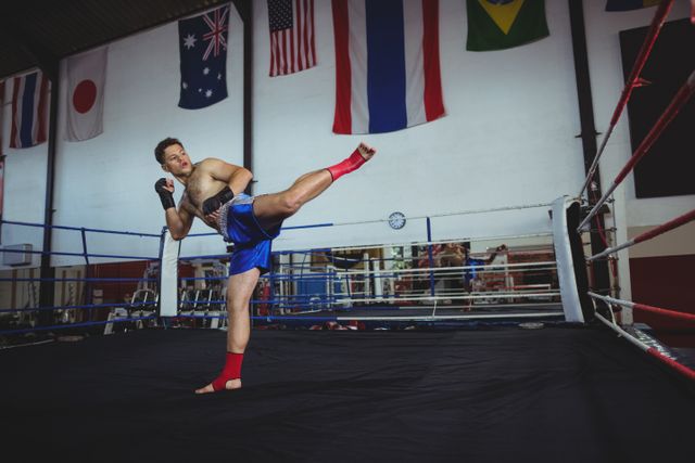 Boxer practicing a high kick in a boxing ring with international flags in the background. Ideal for use in sports and fitness promotions, martial arts training materials, gym advertisements, and motivational content related to athleticism and determination.