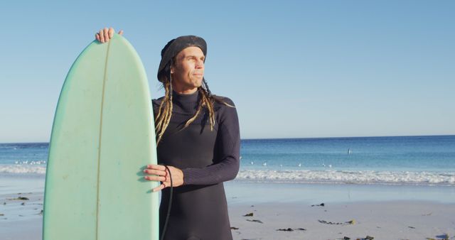 Image of smiling caucasian man with dreadlocks in wetsuit holding surfboard on sunny beach. Freedom, sport, hobbies and healthy active lifestyle concept digitally generated image.