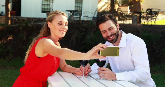 A young Caucasian couple enjoys a moment together taking a selfie with a smartphone at an outdoor setting, with copy space. Their cheerful expressions and wine glasses suggest a casual, romantic outing.