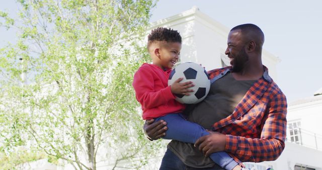 Father holding son as they play with a soccer ball in a backyard on a sunny day. Both are smiling and enjoying quality time together. Suitable for themes related to family bonding, outdoor activities, fatherhood, and active lifestyles. Use for promotions on parenting advice, outdoor sports, and childcare services.
