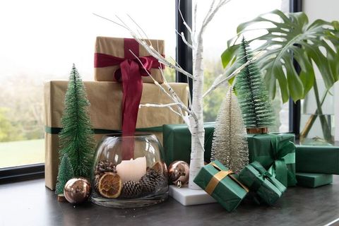 Arranging green and kraft wrapped gifts with bottle-brush trees, candle, monstera leaf