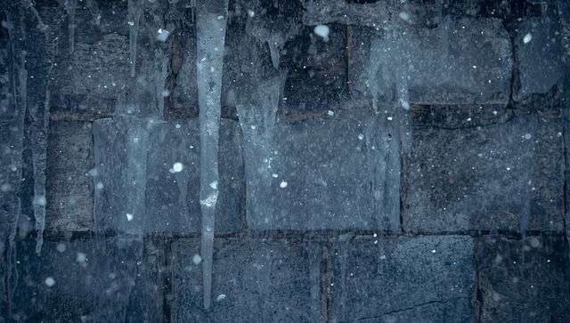 Melting icicles glinting along weathered stone wall with drifting snow and frost