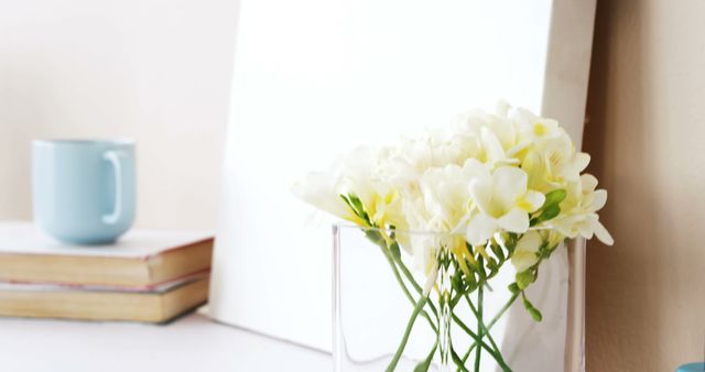 This image showcases white flowers in a clear vase on a table, accompanied by a stack of books and a light blue cup. There is an air of calm and serenity, reflecting a minimalist aesthetic. Perfect for topics related to home decor, minimalism, self-care, and serene interior designs. This is ideal for blogs, websites, or social media content centered on peaceful home settings and decoration ideas.