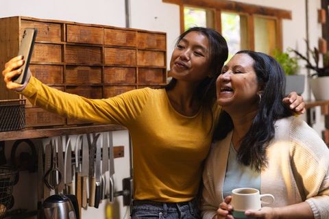 Diverse mother and daughter taking cheerful selfie in warm kitchen
