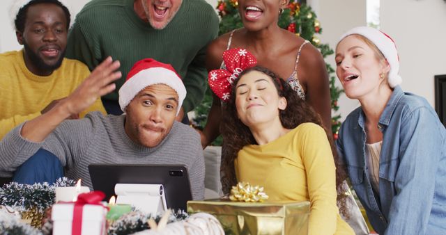 Group of multiethnic family members enjoying Christmas celebration by making a video call. The individuals are wearing festive holiday attire, including Santa hats, and are gathered around a tablet, engaging cheerfully with others remotely. Presents and Christmas decorations provide a festive atmosphere. This could be used for holiday marketing, family celebrations, and social media posts highlighting virtual gatherings and family bonds during festive seasons.