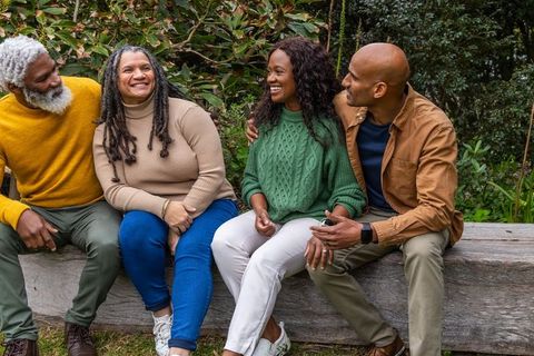 African american friends laughing on log bench in park wearing cozy sweaters and jacket