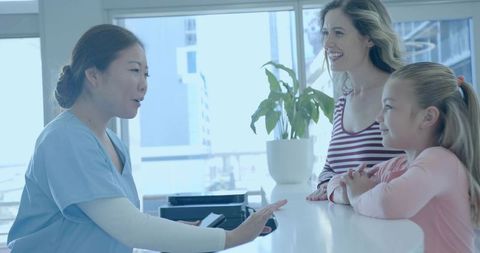 Nurse gesturing while holding documents at reception desk with mother and daughter smiling