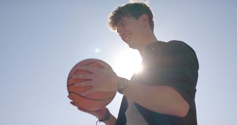Sunlit young man holding basketball smiling under blue sky with lens flare casual fashion