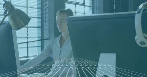 Focused woman typing at multiscreen workstation in modern open-plan office with blinds