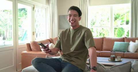 African american man laughing while holding tv remote and relaxing on living room ottoman