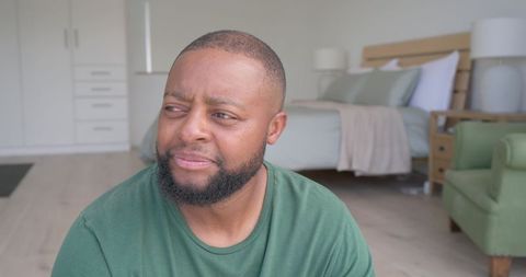 African man sitting in minimalist bedroom wearing green t-shirt looking thoughtful