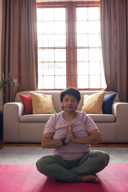 Mature woman sitting cross-legged on a yoga mat with eyes closed, meditating in a cozy living room. Ideal for promoting wellness, mindfulness, and healthy lifestyle. Suitable for articles, blogs, and advertisements related to yoga, meditation, self-care, and active retirement.