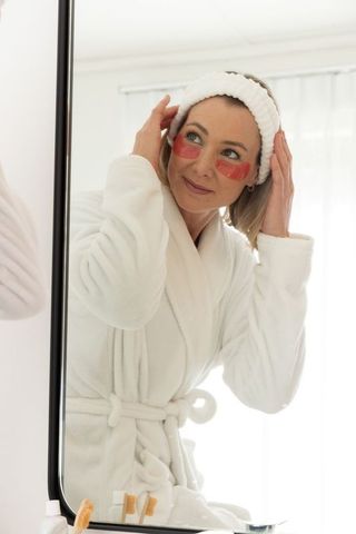 Woman in plush robe adjusting headband with hydrogel eye patches morning skincare routine