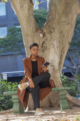 African american woman sitting on urban park bench using smartphone wearing brown coat