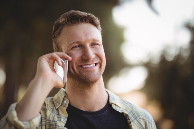 This image shows a young man smiling while talking on his phone outdoors, possibly at an olive farm. It can be used for promoting communication services, lifestyle blogs, or advertisements focusing on rural living and technology.