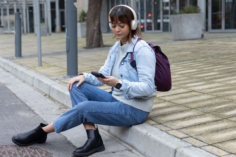 Young woman wearing headphones sitting on curb using smartphone in urban plaza