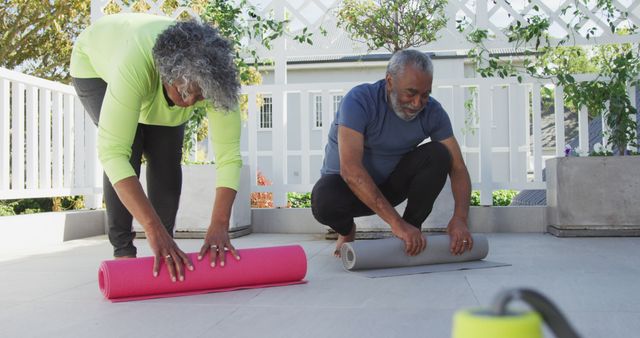 Senior couple is unrolling yoga mats on a sunny terrace for an outdoor exercise routine. They are preparing for a workout in a serene, green environment. This image can be used in articles on elderly fitness, active lifestyles for seniors, yoga and wellness practices, or outdoor recreational activities.