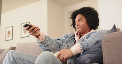 African american man relaxing on sofa watching tv with remote and popcorn in denim jacket