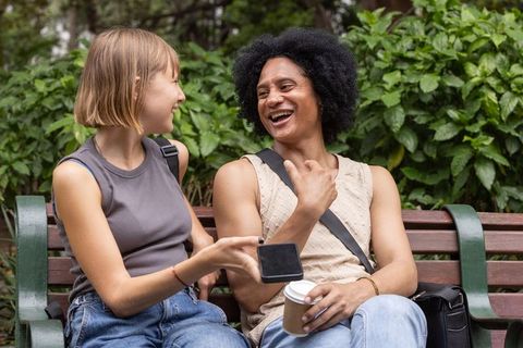 Laughing diverse friends sharing smartphone and coffee on park bench in sunny urban park
