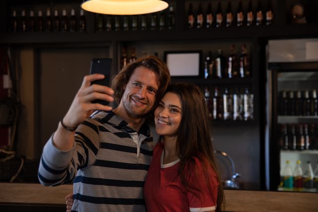 Group of multi-ethnic male and female friends at the bar in a pub during the day, one woman holding a smartphone and taking a selfie of all of them smiling and laughing together. Friendship leisure time fun.