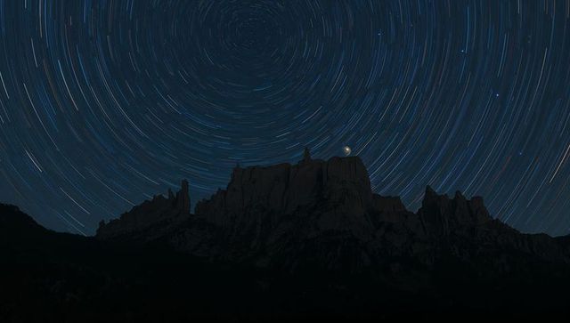 Circling star trails over jagged mountain silhouette with crescent moon on rock spire