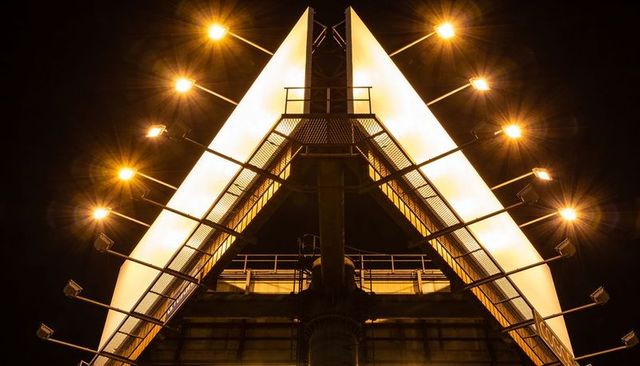 Illuminated twin-triangle bridge pylon at night with grated walkway and starburst lamps