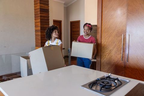 Diverse female friends moving into modern kitchen carrying cardboard boxes and smiling