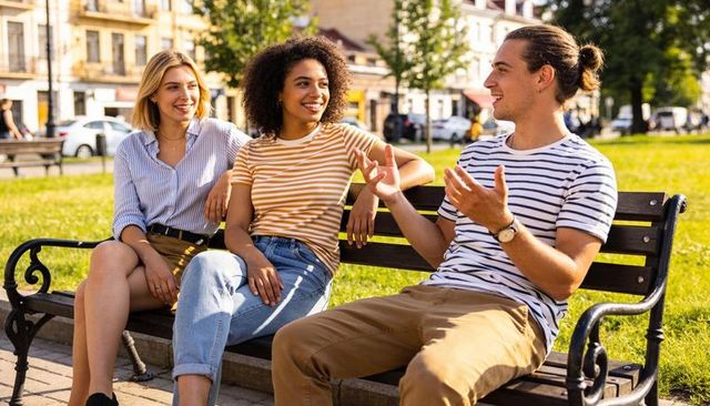 Three friends chatting on park bench, diverse young adults relaxing in sunny urban park