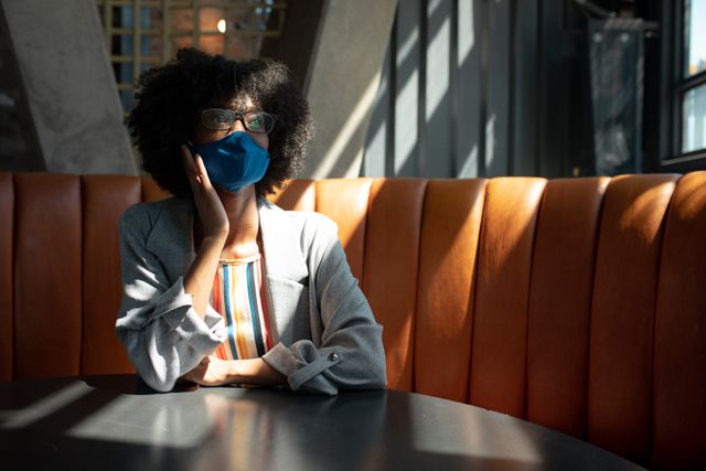 African american woman wearing face mask sitting in cafe looking out of window. out and about in the city during covid 19 pandemic.