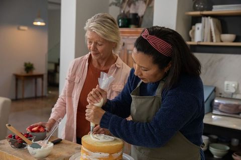 Diverse women frosting layered sponge cake together in cozy kitchen, senior woman guiding