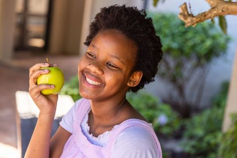 African american teen girl smiling holding green apple on sunlit porch in pink pinafore