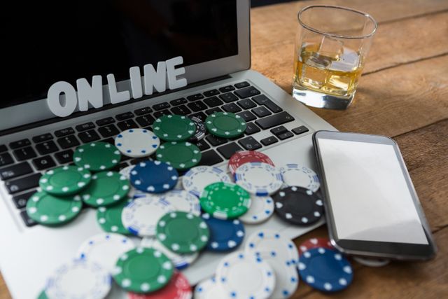Image depicts a laptop with the word 'ONLINE' and scattered poker chips, a smartphone, and a glass of whiskey on a wooden table. Ideal for illustrating concepts related to online gambling, digital betting, and casino gaming. Can be used in articles, blogs, and advertisements focusing on online casinos, gambling addiction, or digital entertainment.