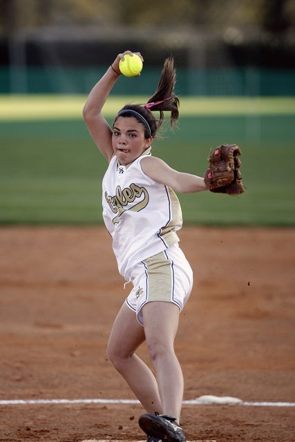 Caucasian female pitcher in mid-action, throwing a ball during a competitive softball game on an outdoor field. Perfect for content related to sports, fitness, female athletes, women's competitions, and team sports activities. Ideal for marketing materials, articles about athletics, and fitness promotional content.