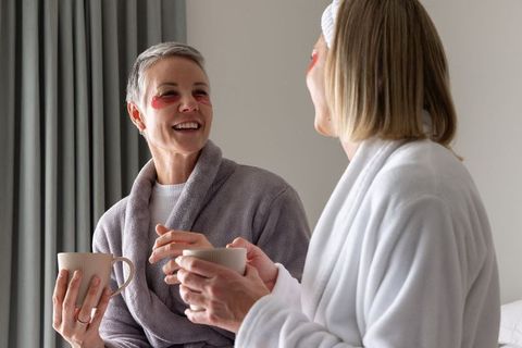 Senior woman and friend sharing morning skincare ritual in robes with eye patches and coffee