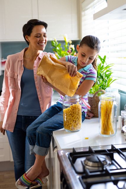 Vertical of smiling caucasian mother and daughter in kitchen, girl pouring pasta in jar, copy space. Family, childhood, domestic life and happiness concept.