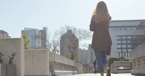 Backlit young woman walking through urban plaza with concrete planters and skyscrapers