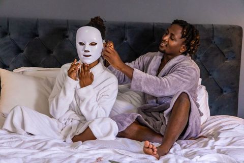 African american couple relaxing on bed in plush robes while man adjusting led facial mask