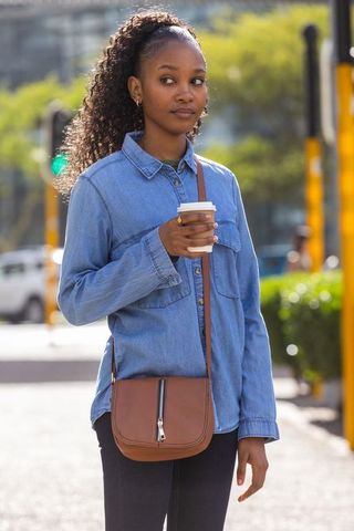 African woman holding coffee wearing denim shirt and crossbody leather bag on city sidewalk