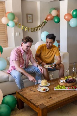 Two men celebrating birthday at home cutting cake on sofa surrounded by balloons
