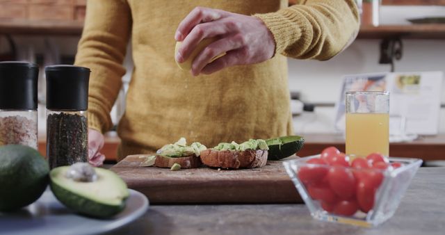 Man in mustard sweater preparing avocado toast while squeezing lemon over it in a modern kitchen. Fresh ingredients like avocados and cherry tomatoes are on the table with a glass of juice. Ideal for use in food blogs, healthy diet promotion, breakfast recipes, and kitchen appliance advertisements.