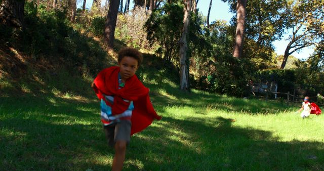 A young African American boy is running energetically in a park, with a child in a red cape visible in the background. Their playful activity suggests a superhero game or imaginative outdoor playtime.